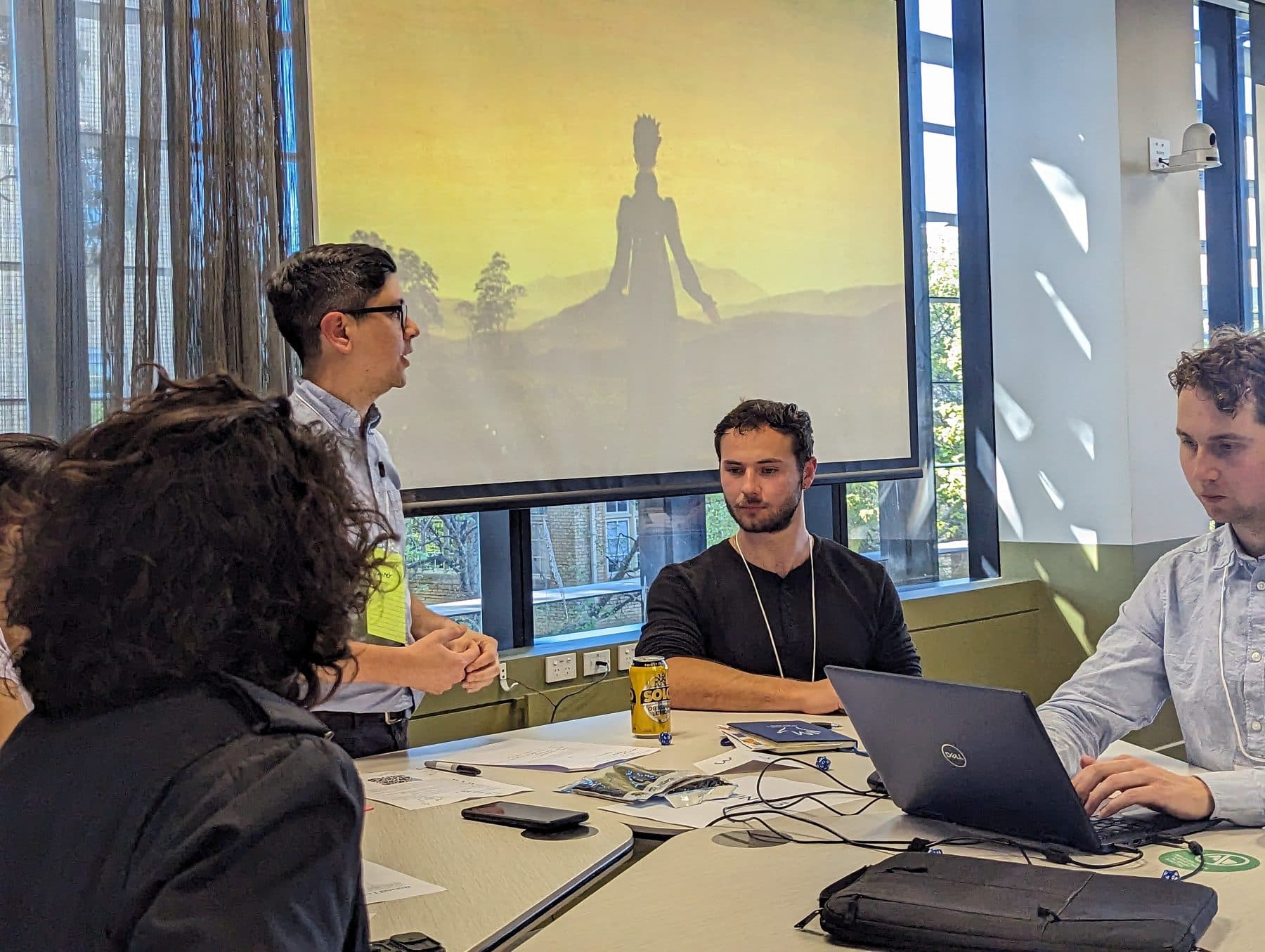 Players negotiate around a table at the Before the Rising Sun wargame, the game's golden sunset scenario slide glowing on the wall behind them.